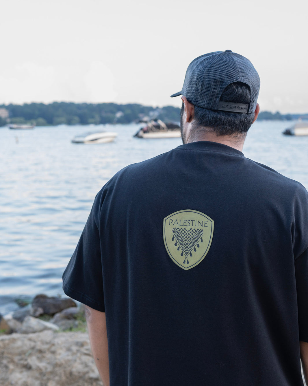 Person wearing a black t-shirt with 'Palestine' logo by a body of water