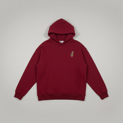 A red pullover hoodie with a small embroidered logo on the left chest, laid flat on a white sheet. The bottom of a person's legs wearing gray sweatpants and leopard-print slippers are visible at the bottom edge.